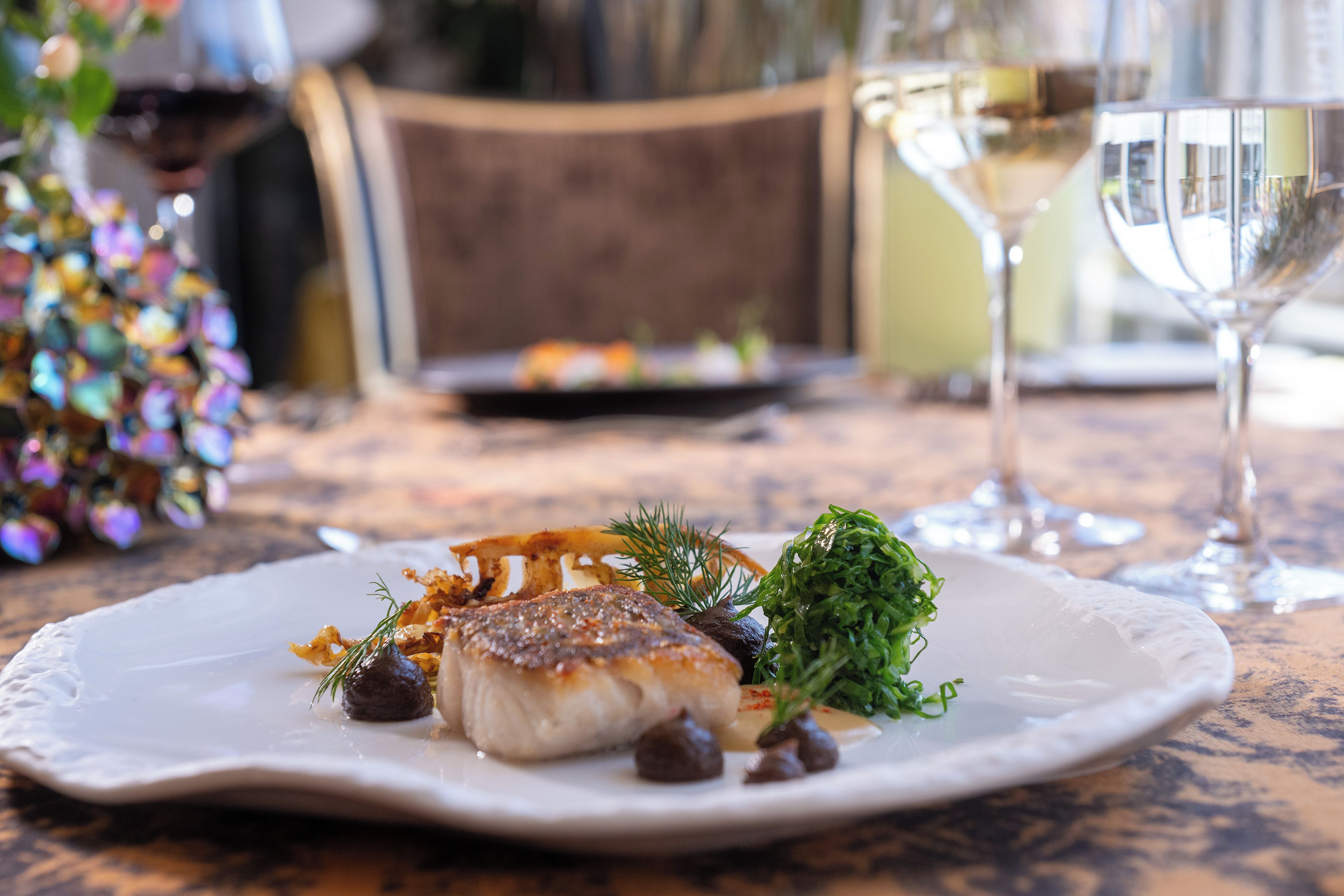 A plate of food with a white plate, a glass of white wine, and a chair in the background.