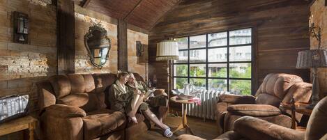 A rustic cabin interior with leather sofas, a stone fireplace, and a large window with a view of greenery.