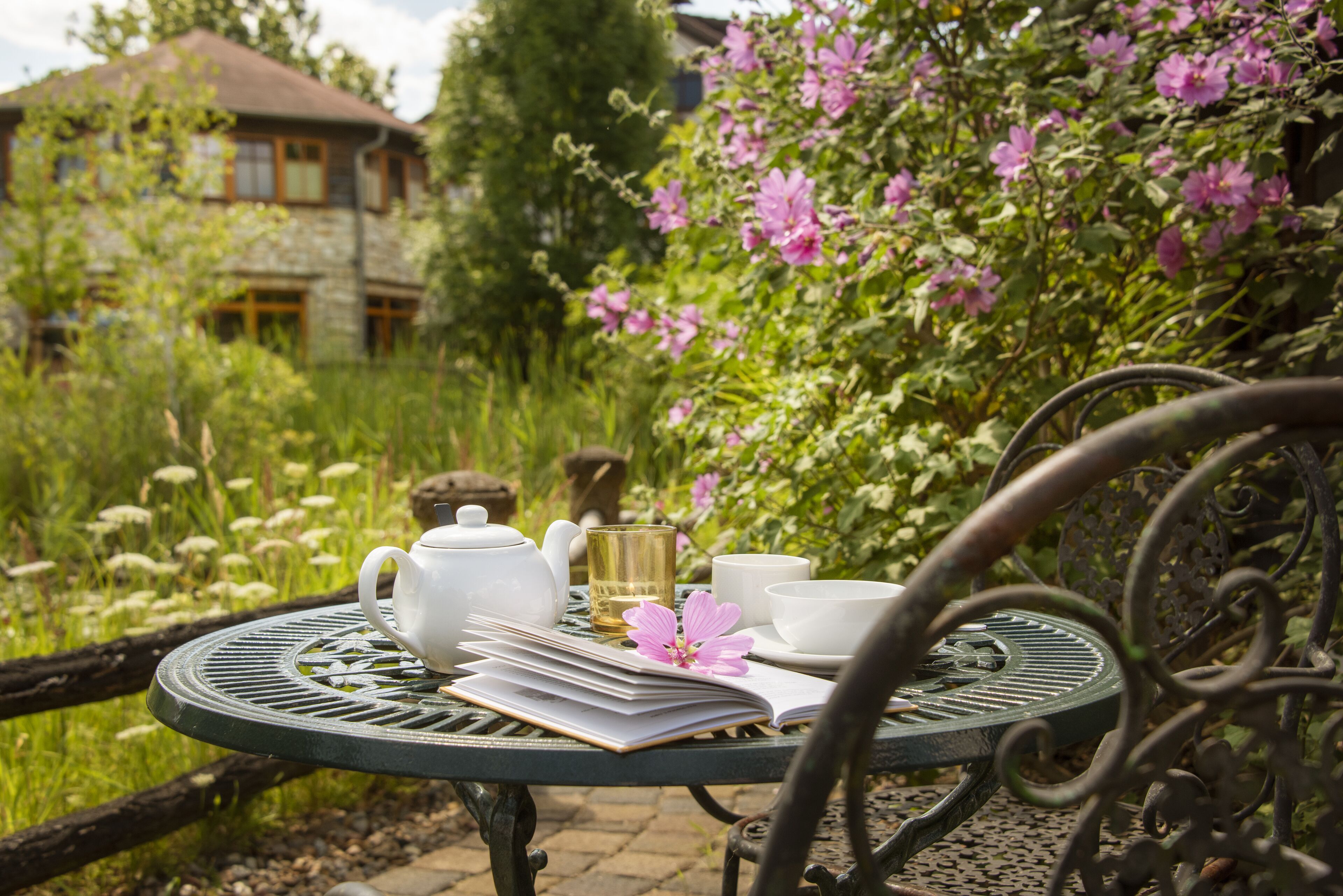 Una mesa de jardín con una tetera, tazas y un libro, rodeada de abundante follaje y flores.