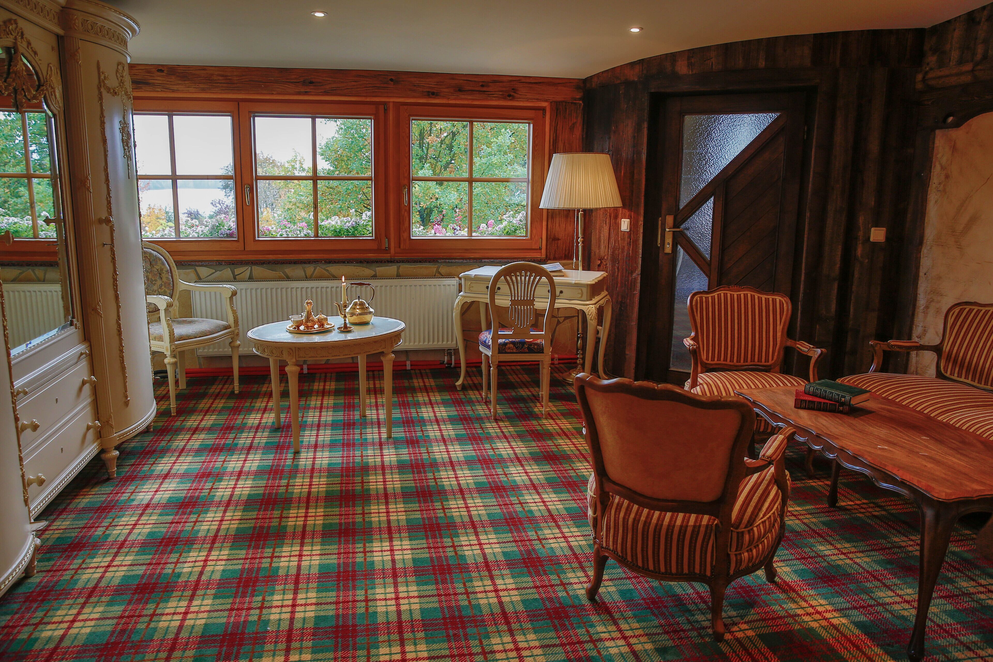 Une pièce dotée d’un tapis à carreaux, de meubles en bois, d’une table avec des chaises, d’un radiateur et de grandes fenêtres donnant sur un paysage verdoyant.
