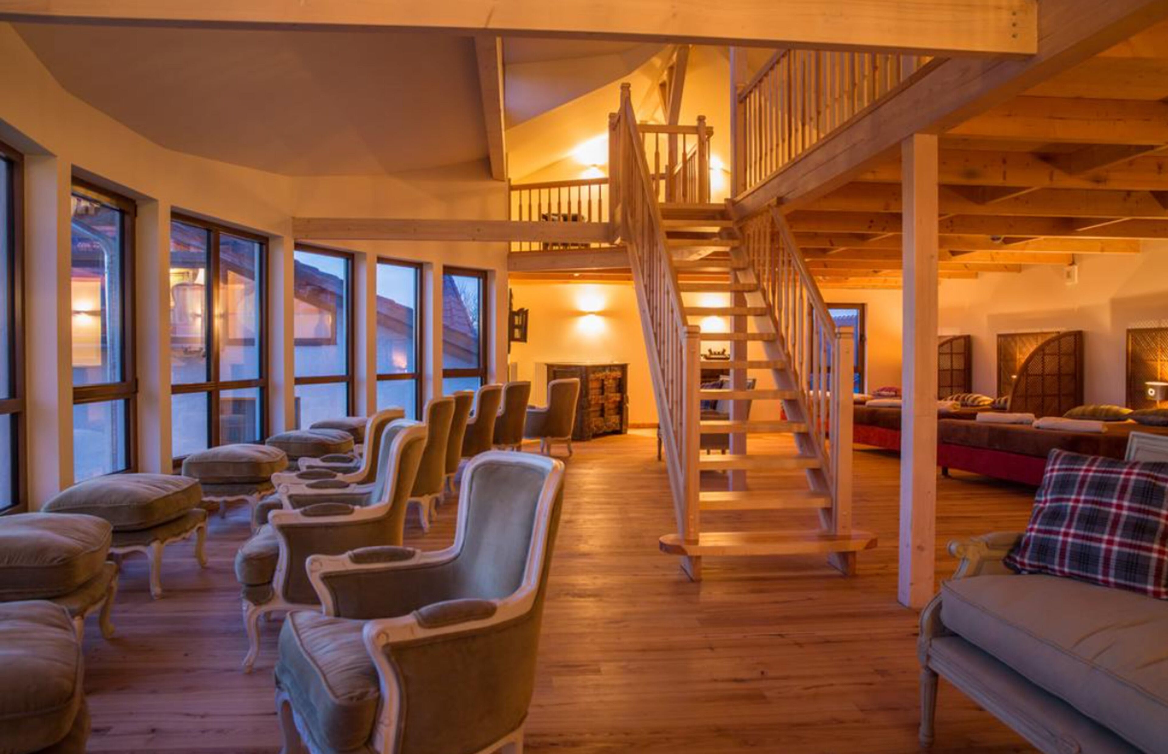 Una habitación amplia con una escalera de madera, varios espacios de descanso y grandes ventanales.