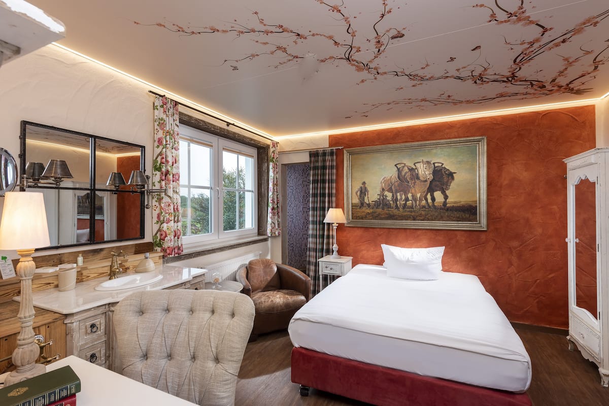 Ein Hotelzimmer mit Bett, Sessel, Schreibtisch mit Lampe, Spiegel, einem Bild an der Wand und einem Fenster mit Vorhängen.