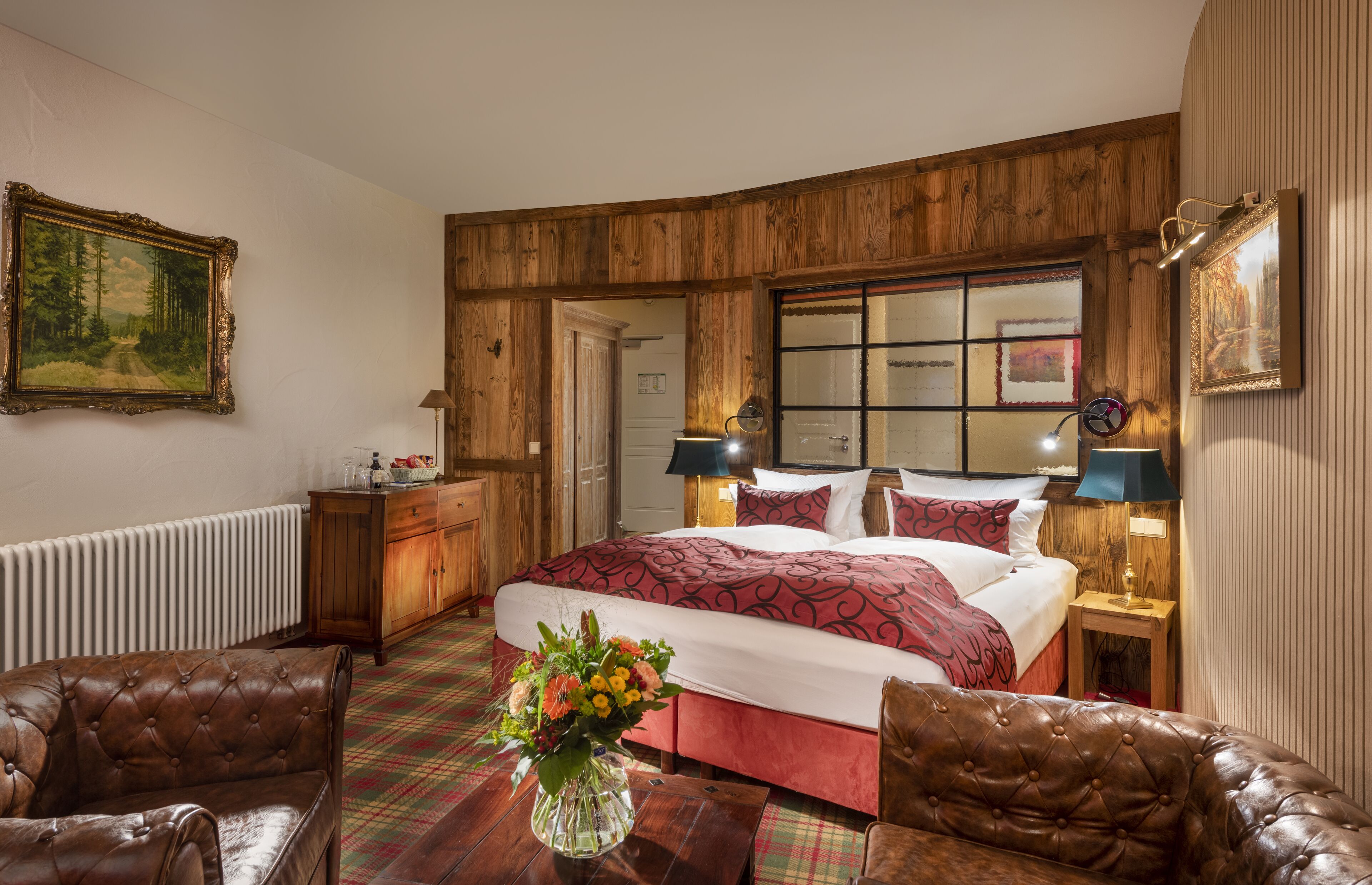 A bedroom with a large bed, wooden paneling, a radiator, a dresser, two lamps, and a vase with flowers.
