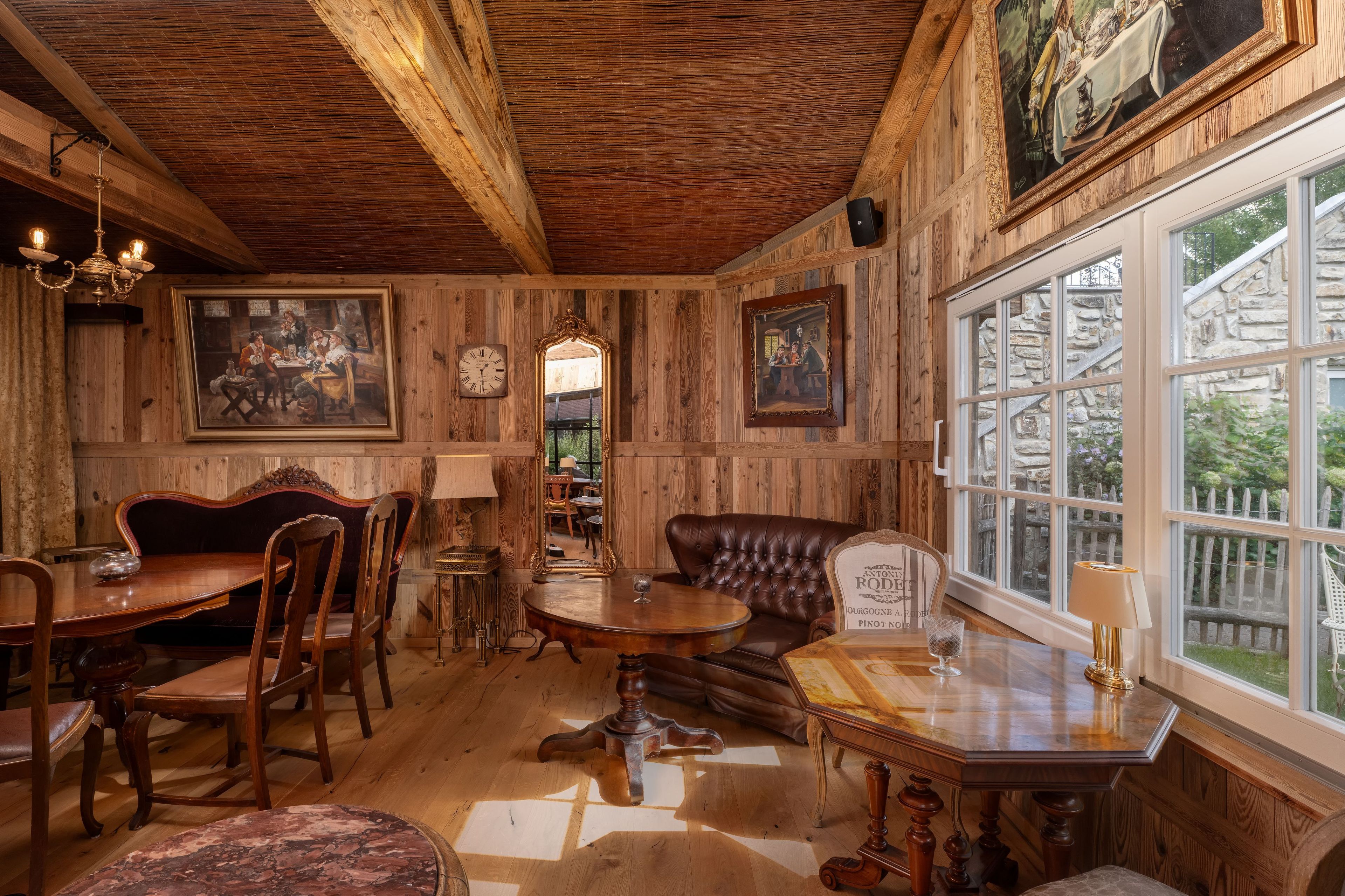 Un interior rústico con paredes de madera, una zona de comedor con mesa y sillas de madera, un sofá de cuero y un ventanal con vistas a la vegetación.