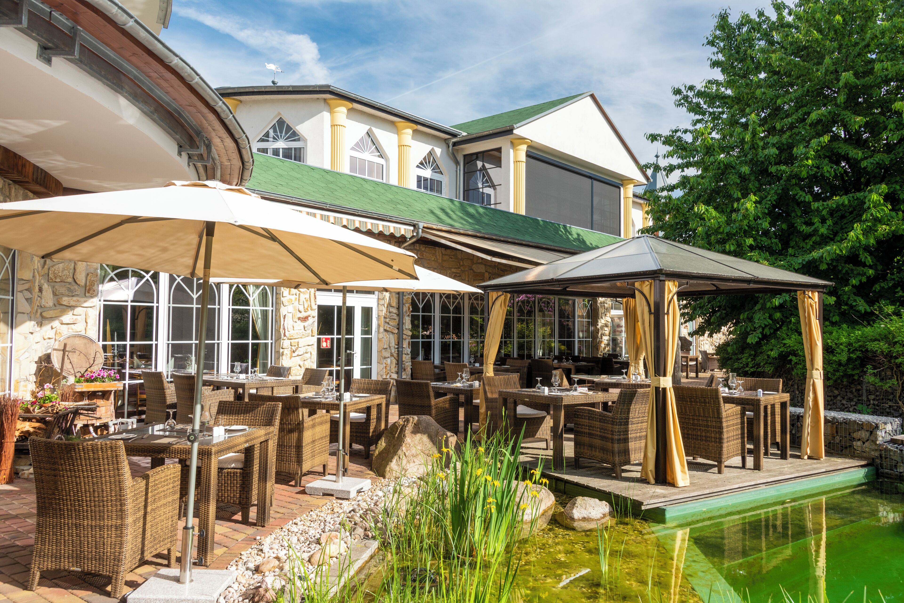 Ein Außenbereich zum Essen mit Korbmöbeln und Sonnenschirmen, ein Steinhaus mit großen Fenstern und ein kleiner Teich.