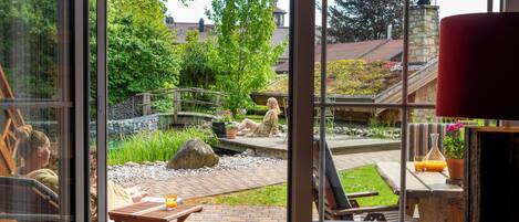 A view from a window of a patio area with a wooden table, a bench, and a small bridge over a garden.