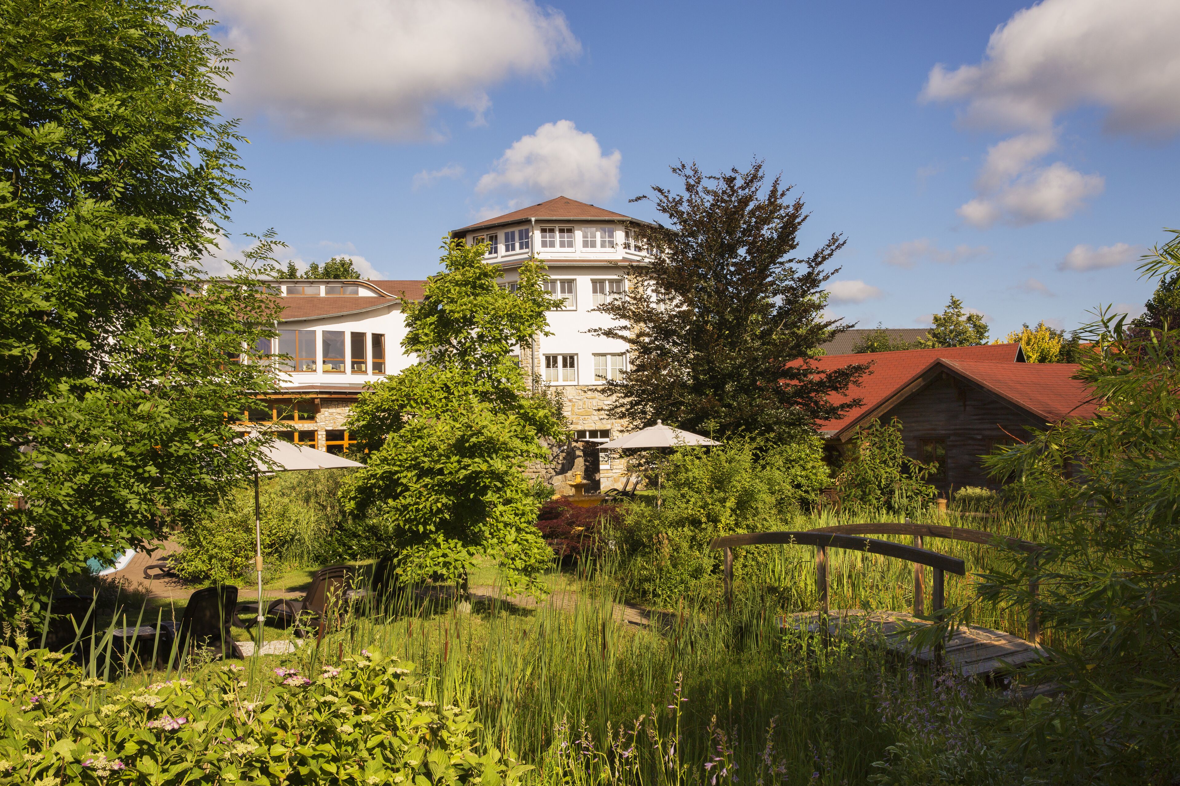 A large, multi-story building with a red roof, surrounded by lush greenery and a wooden bridge.