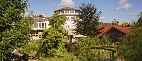 A large, multi-story building with a red roof, surrounded by lush greenery and a wooden bridge.