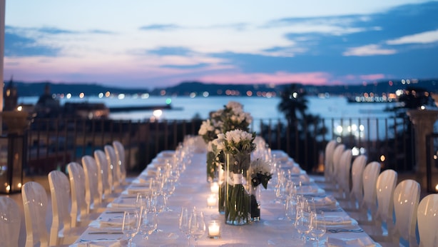 Una larga mesa de comedor dispuesta para un evento formal, con sillas a ambos lados, un centro de mesa floral y vistas al agua y las luces de la ciudad al fondo.