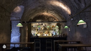 Un bar con un arco de piedra rústica, luces colgantes verdes tipo farolillos y una variedad de botellas en estantes.