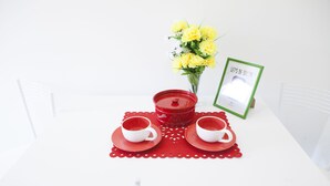 Una mesa blanca con dos tazas blancas sobre platillos rojos, una tetera roja y un ramo de flores amarillas.