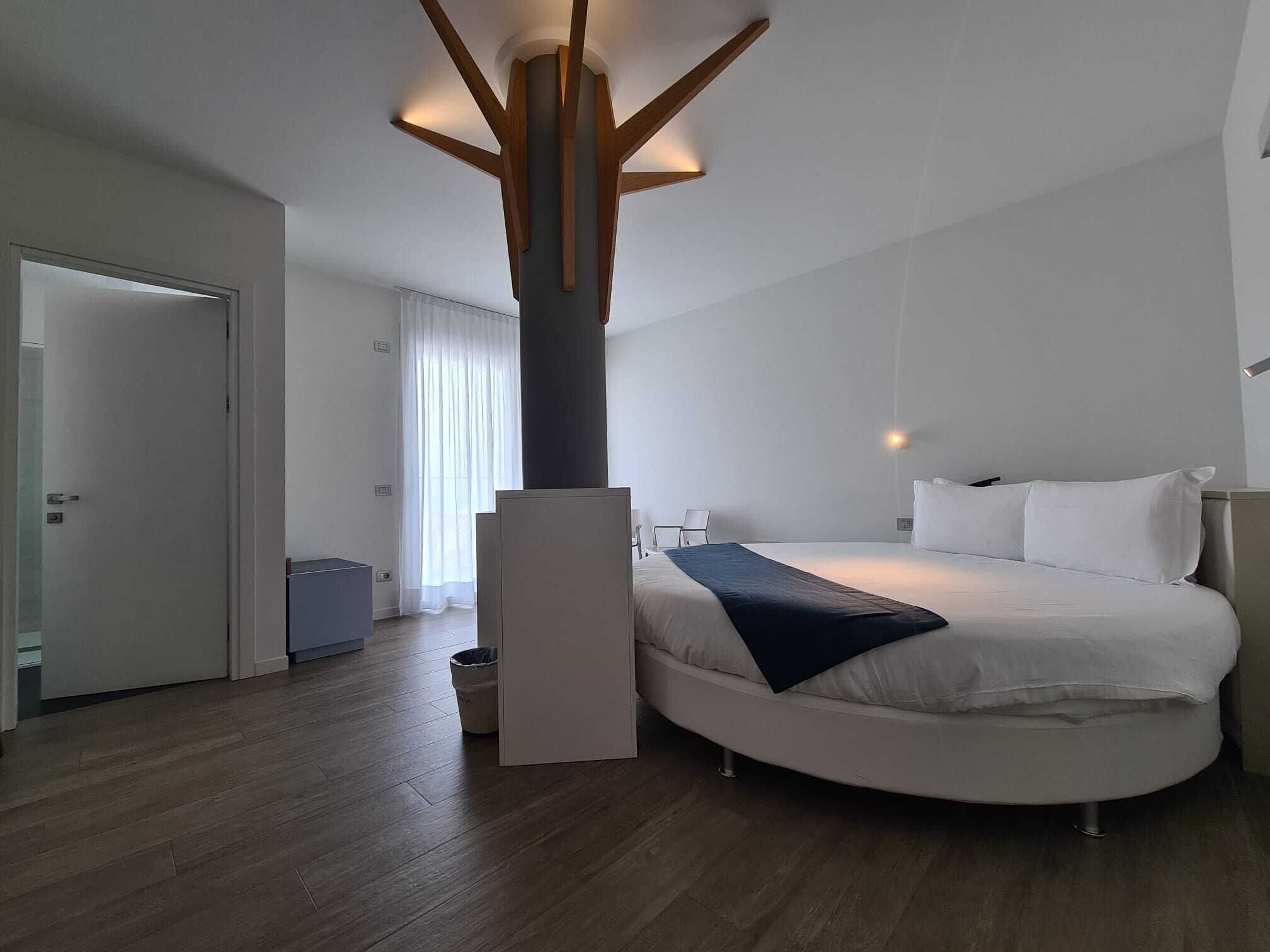 A modern hotel room with a large bed, a wooden ceiling structure, and a view of the outside through a window.