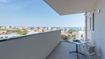 Ein Balkon mit Fliesenboden, einem Tisch und zwei Stühlen. Blick auf eine Stadtlandschaft mit Gebäuden und blauem Himmel.