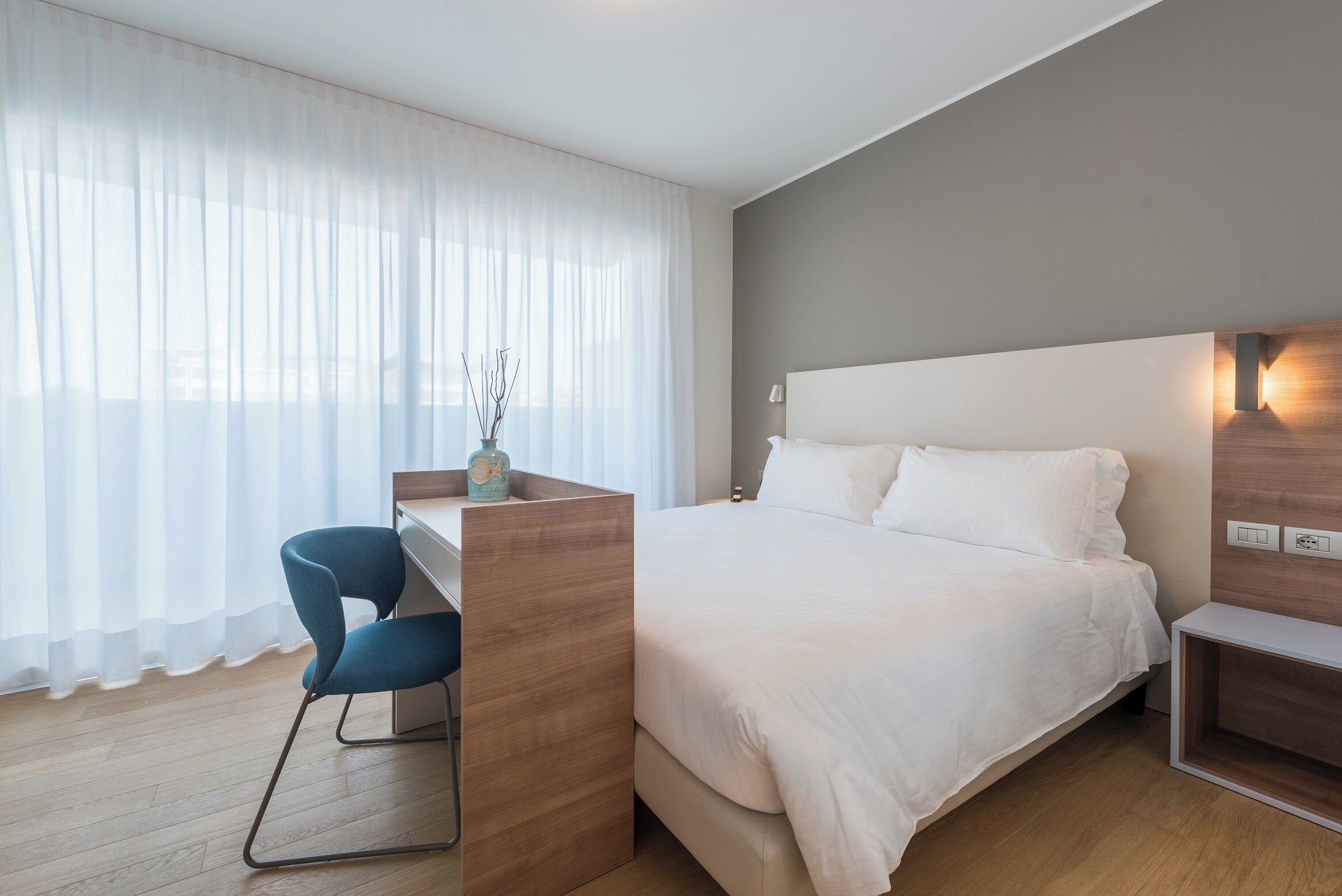 A modern hotel room with a bed, a desk with a chair, and a large window with sheer curtains.