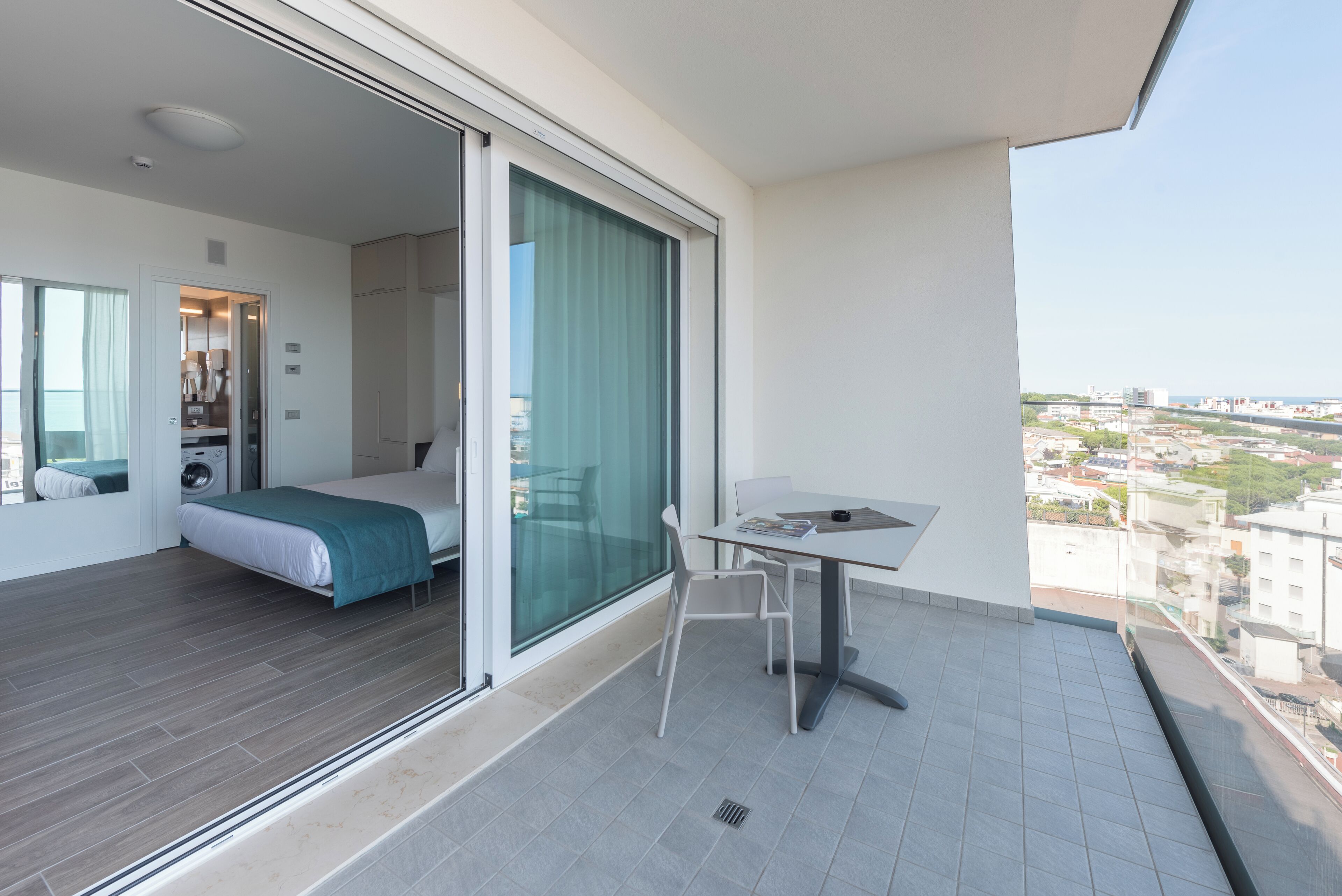 A modern hotel room with a balcony, a bed, a table, and a view of the city.