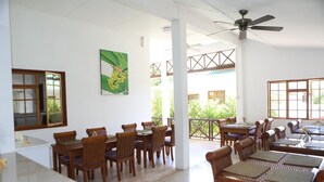 Une salle à manger spacieuse, dotée de tables et de chaises en bois, d’un ventilateur de plafond et de grandes fenêtres offrant une vue sur la verdure environnante.