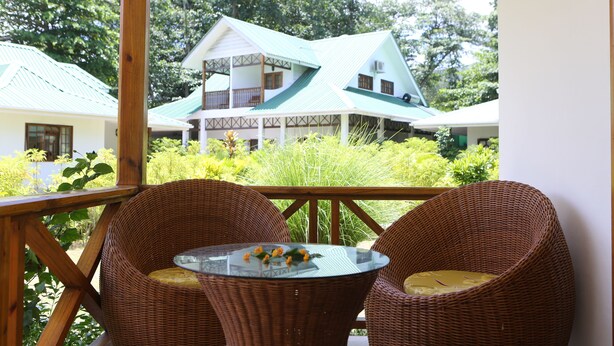 Un balcon avec des fauteuils en osier et une table en verre, donnant sur une maison avec un toit végétalisé et des murs blancs.