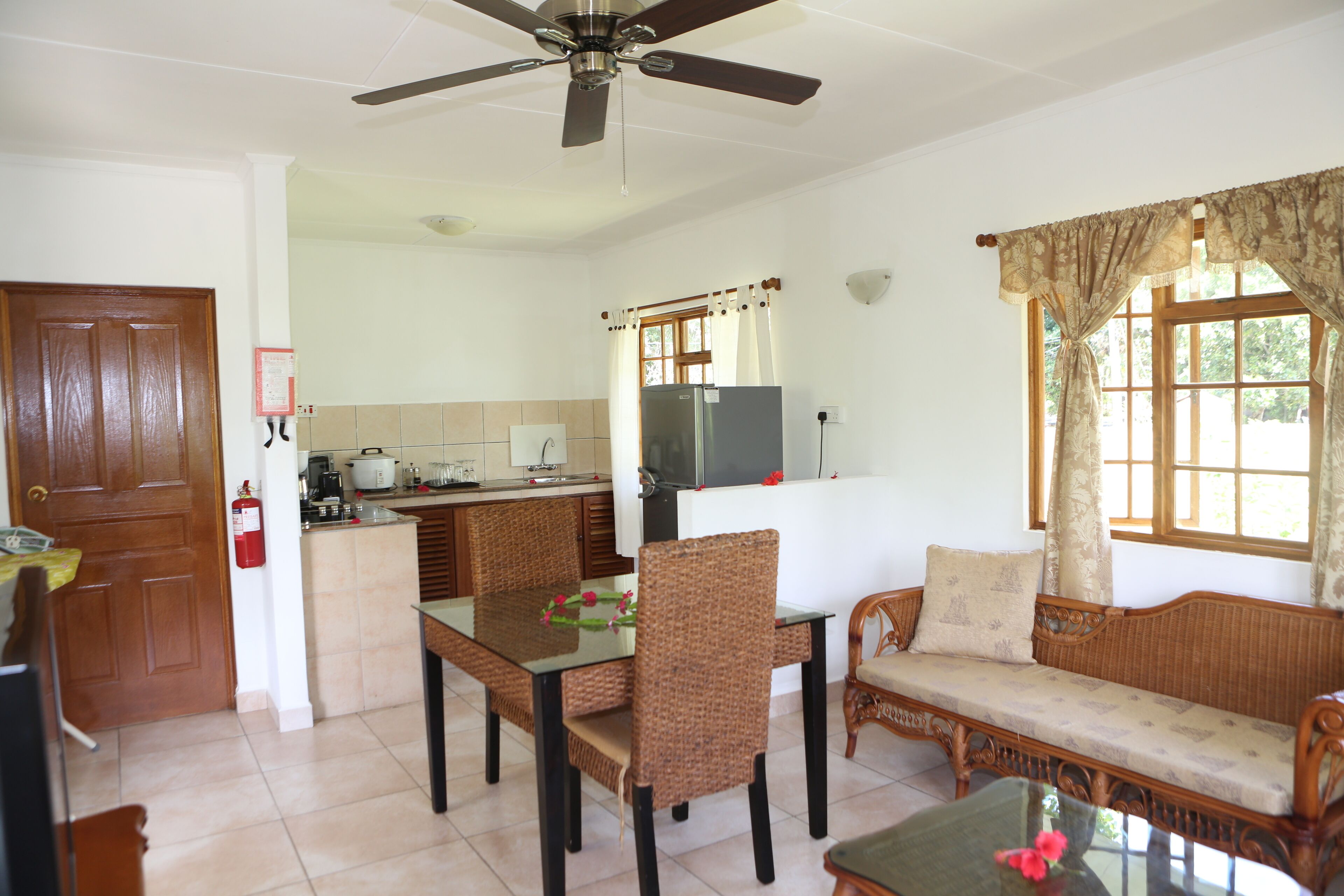Une pièce comprenant une table à manger, des chaises, un canapé, un ventilateur de plafond, un coin cuisine et une fenêtre avec des rideaux.
