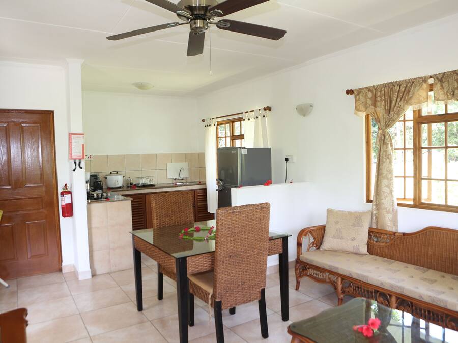 A room with a dining table, chairs, a sofa, a ceiling fan, a kitchen area, and a window with curtains.