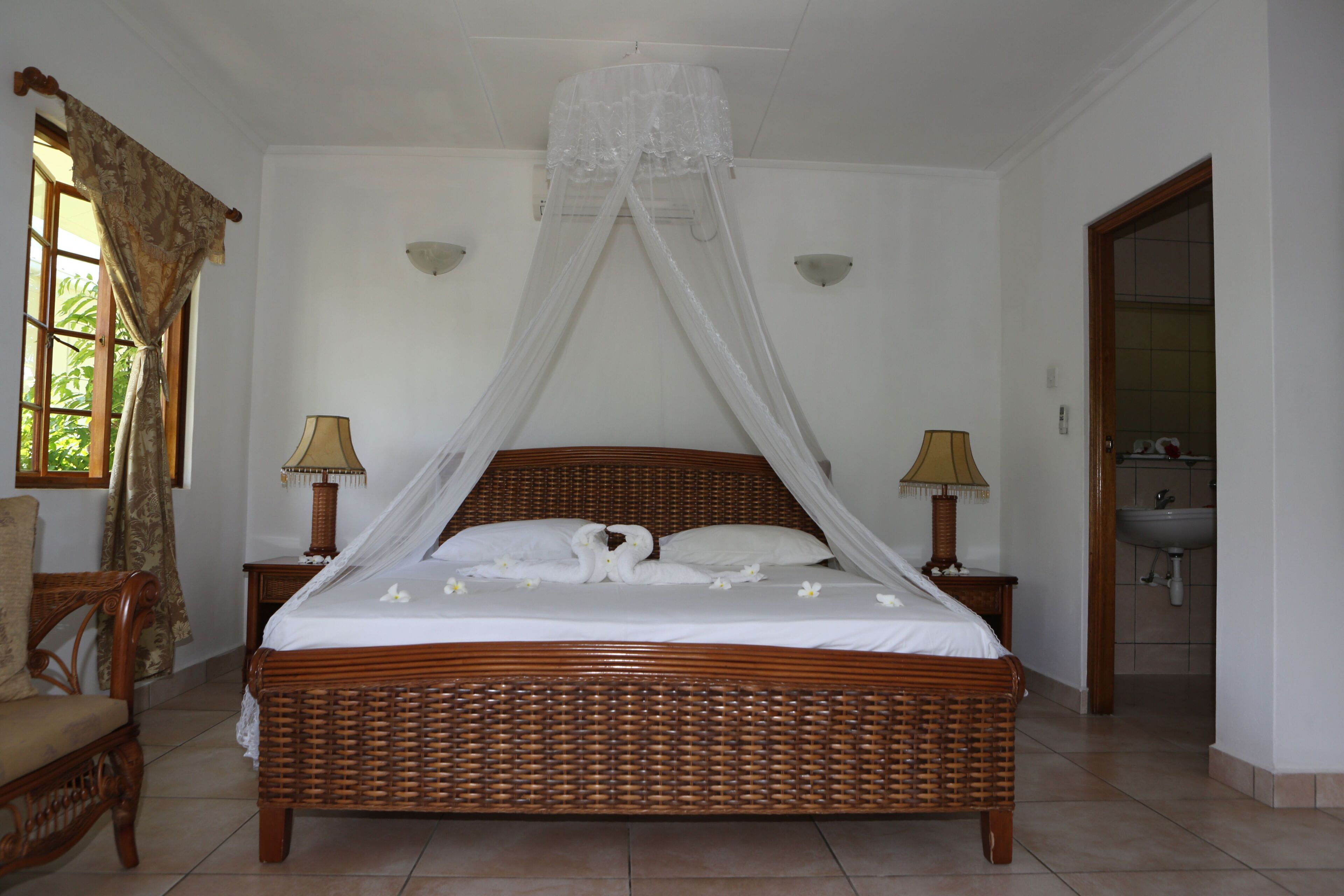 Un dormitorio amplio con una cama grande, dos mesitas de noche, un dosel sobre la cama, una silla y un baño visible a través de una puerta abierta.