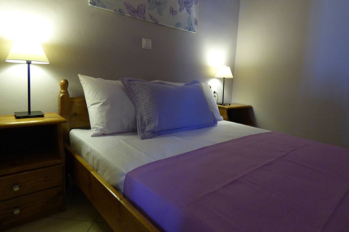 A neatly made bed with a purple bedspread, two white pillows, and two bedside lamps on wooden nightstands.