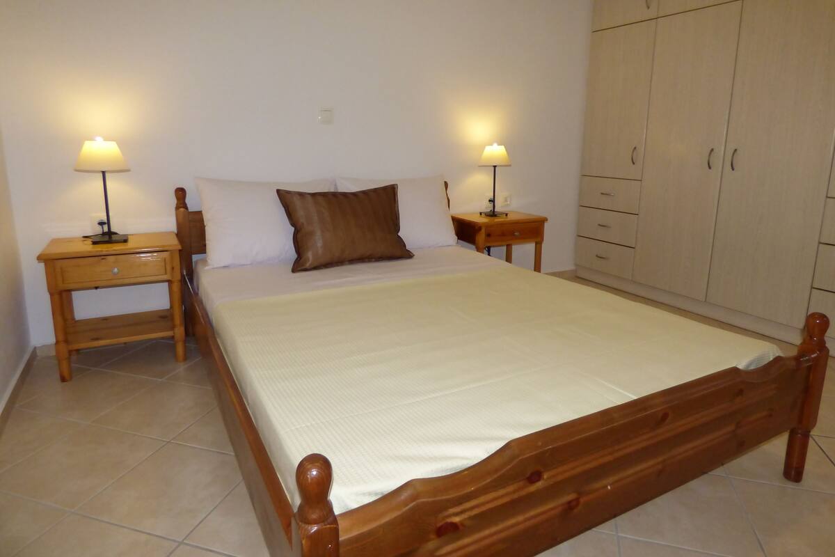 A bedroom with a wooden bed, two bedside tables with lamps, a wardrobe, and a tiled floor.