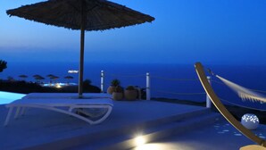 A poolside area with a lounge chair, a thatched umbrella, and a hammock. - Rampelia apartments (Santorini)