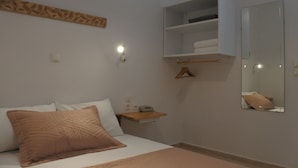 A bedroom with a bed, a bedside table, a mirror, and a shelf. - Rampelia apartments (Santorini)