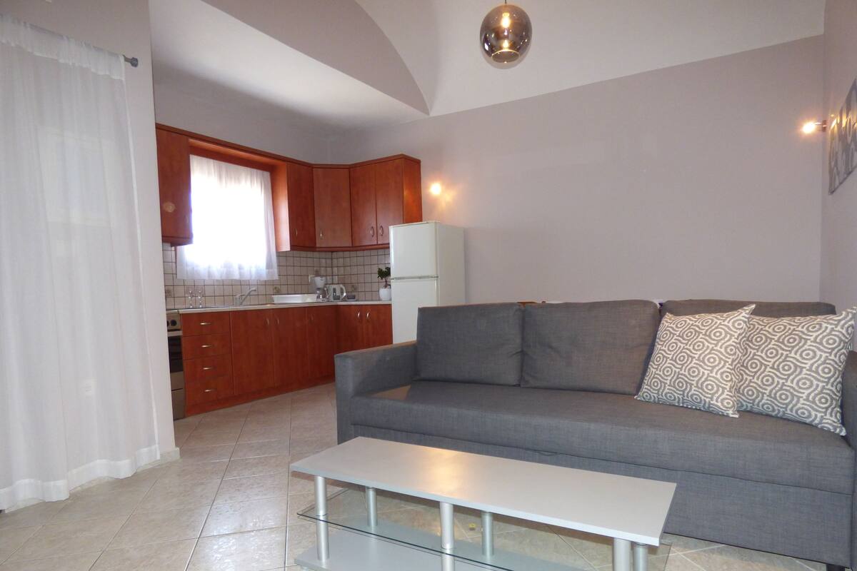 A living room with a grey sofa, patterned cushions, a glass coffee table, and a kitchen in the background.