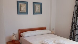 A neatly made bed with white linens, a wooden nightstand with a lamp, and two framed pictures on the wall. - Apartamentos Danesp - Marina D'Or Primera Línea (Oropesa del Mar)