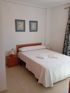 A neatly made bed with white linens, a wooden nightstand with a lamp, and two framed pictures on the wall. - Apartamentos Danesp - Marina D'Or Primera Línea (Oropesa del Mar)