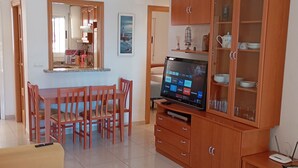 A living room with a sofa, TV, wooden cabinet, dining area with chairs, and a kitchen in the background. - Apartamentos Danesp - Marina D'Or Primera Línea (Oropesa del Mar)