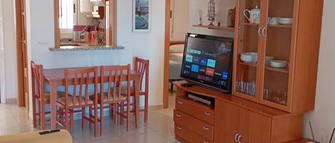 A living room with a sofa, TV, wooden cabinet, dining area with chairs, and a kitchen in the background.