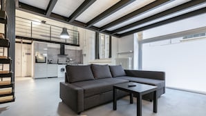 A modern living room with a grey sofa and a small table, a kitchen in the background, and a large window providing natural light.