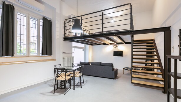 A spacious living area with a loft bedroom, a dining table with chairs, a grey sofa, and a staircase with a wooden finish.