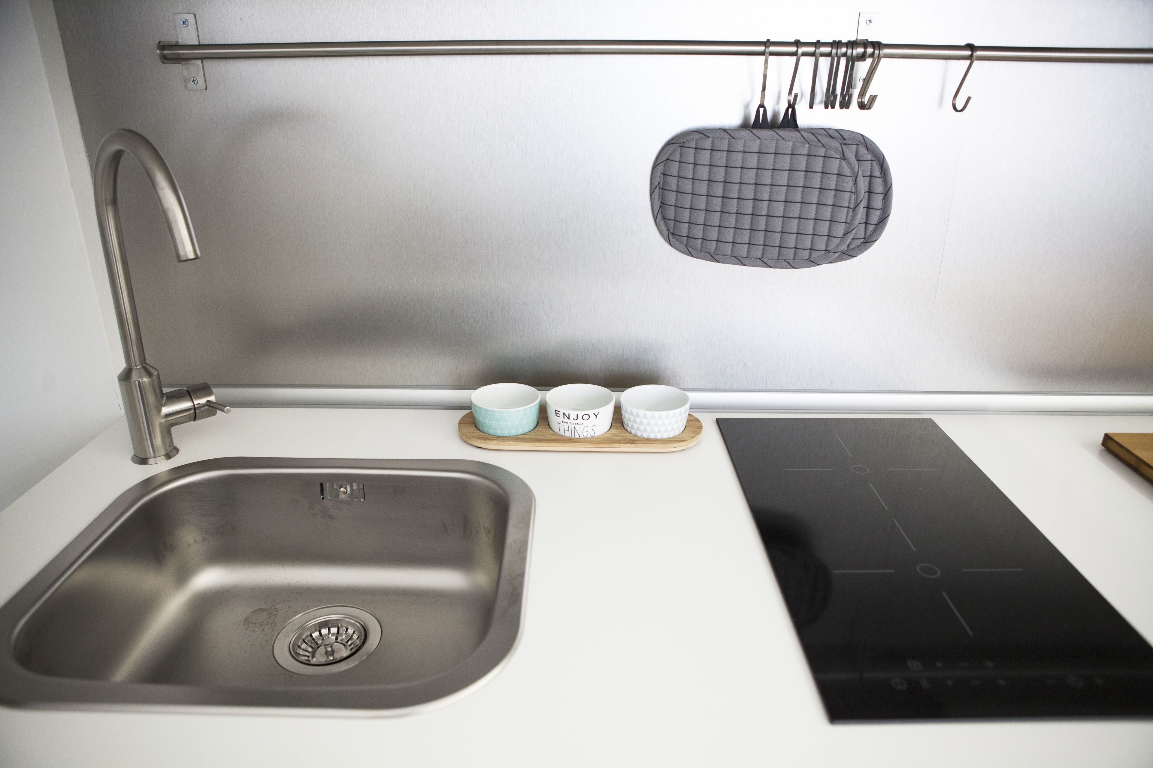 A kitchen with a stainless steel sink, a black induction cooktop, and three ceramic cups on a wooden tray.
