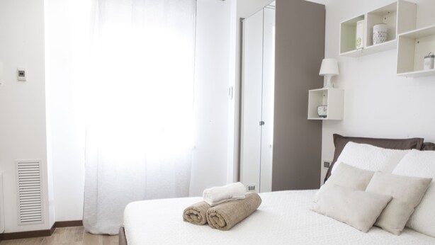 A neatly made bed with rolled towels, a bedside table with a lamp, and a shelf with decorative items. - Alessia's Flat - Paolo Sarpi (Milan)