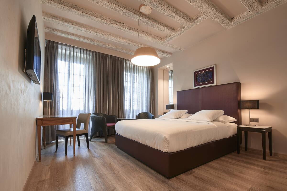 A hotel room with a large bed, a desk, a chair, a TV, and a window with curtains.