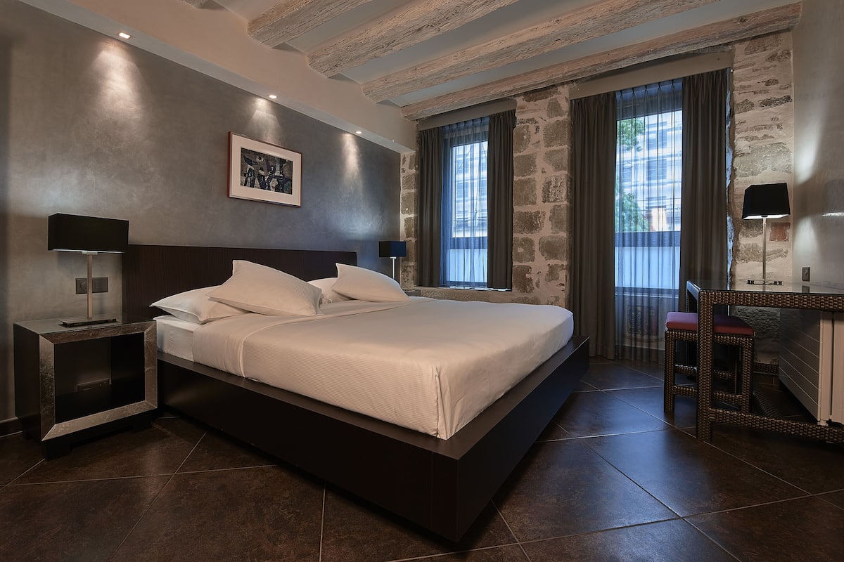 A modern bedroom with a large bed, a bedside table, a lamp, and a view of the outside through a window.