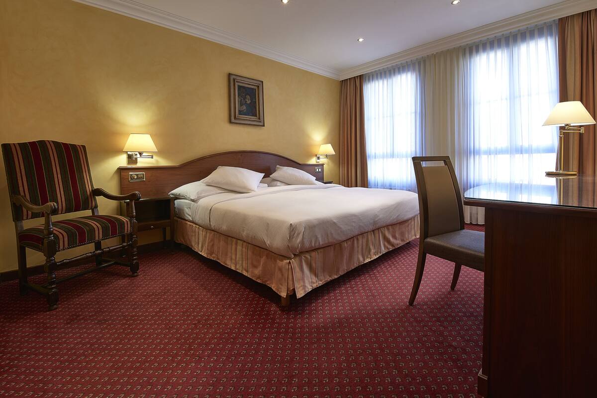 A hotel room with a large bed, two bedside lamps, a wooden chair, a desk, and a large window with curtains.
