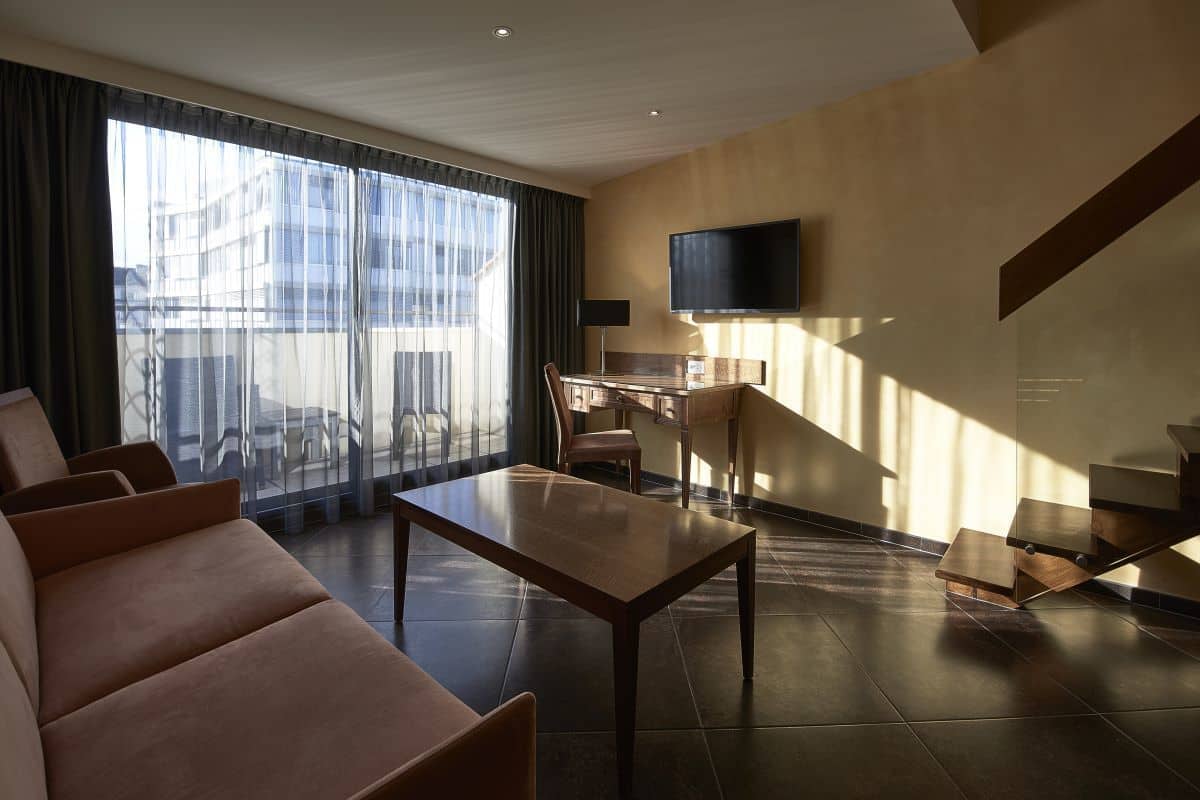 A modern hotel room with a large window, a flat-screen TV, a sofa, a dining table, and a staircase.