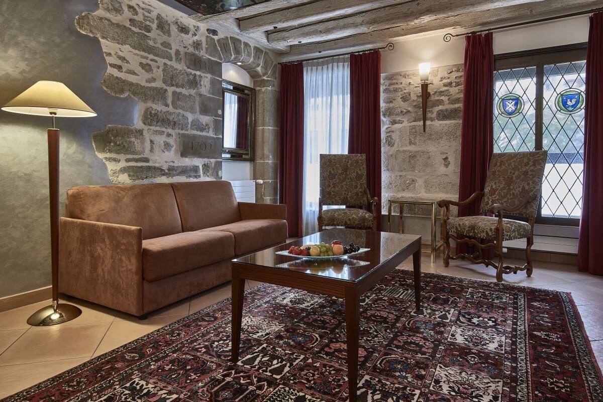 A room with a stone wall, a brown sofa, a glass coffee table with fruit, patterned chairs, and a large rug.