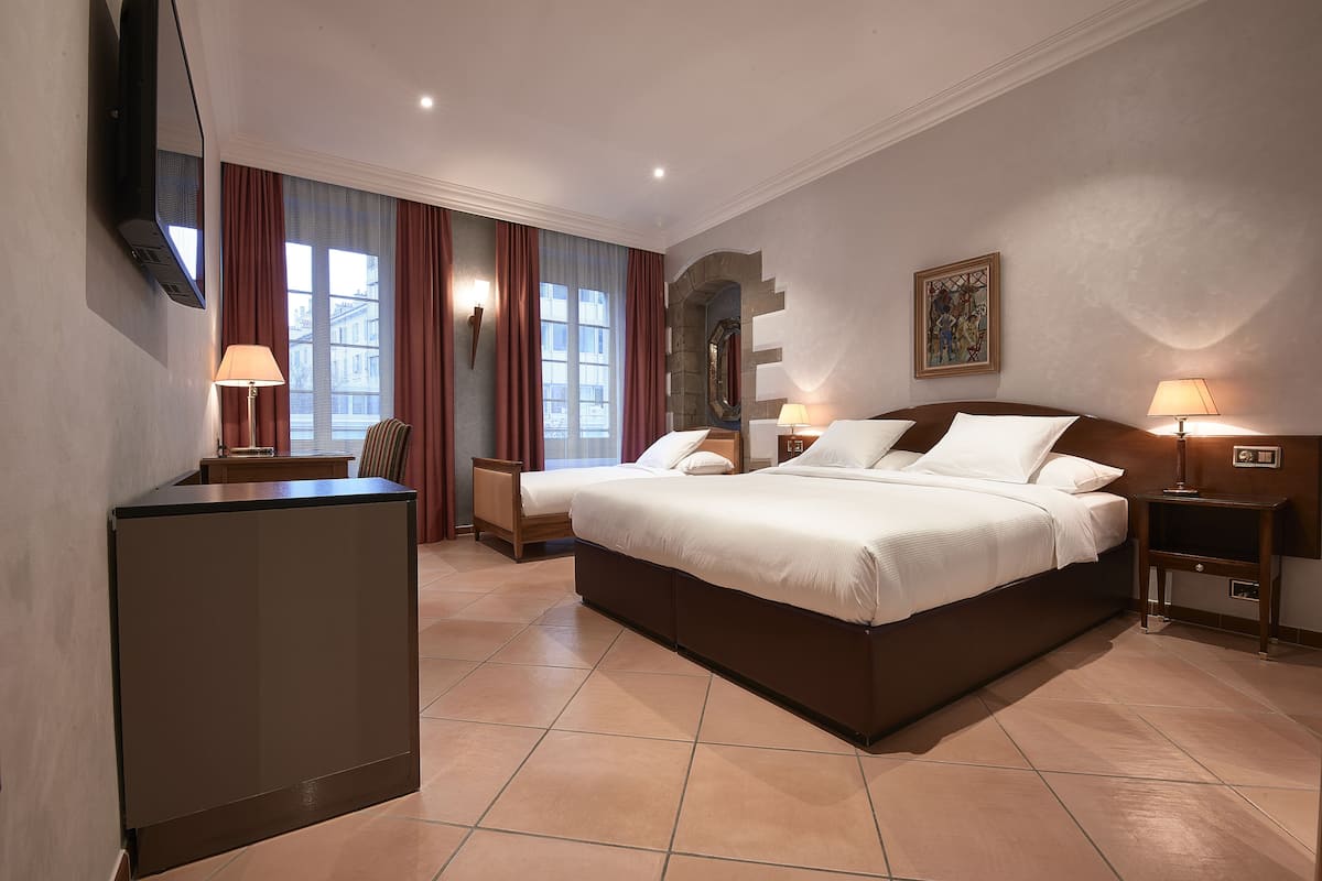 A hotel room with a large bed, two bedside tables, a desk, a TV, and a large window with curtains.