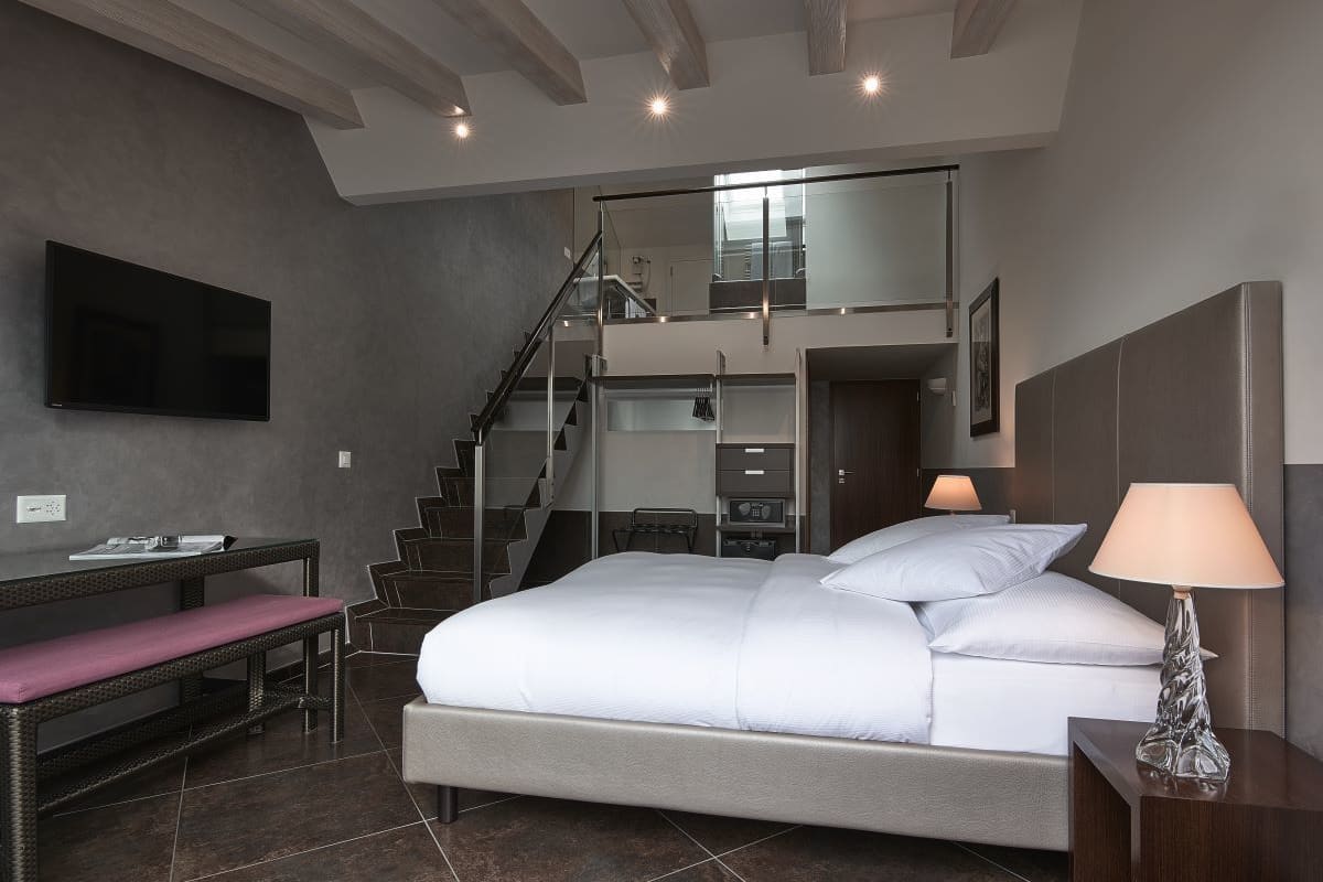 A modern hotel room with a large bed, a desk, a bench, and a TV.