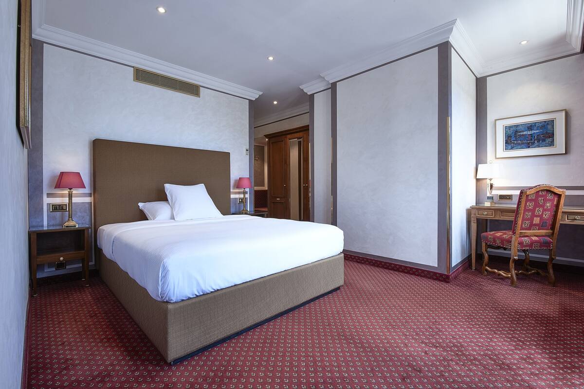 A hotel room with a large bed, a desk with a chair, a nightstand, and a lamp.