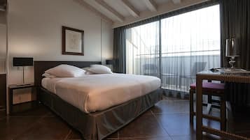 A hotel room with a large bed, a desk with a chair, a window with curtains, and a wall-mounted lamp.