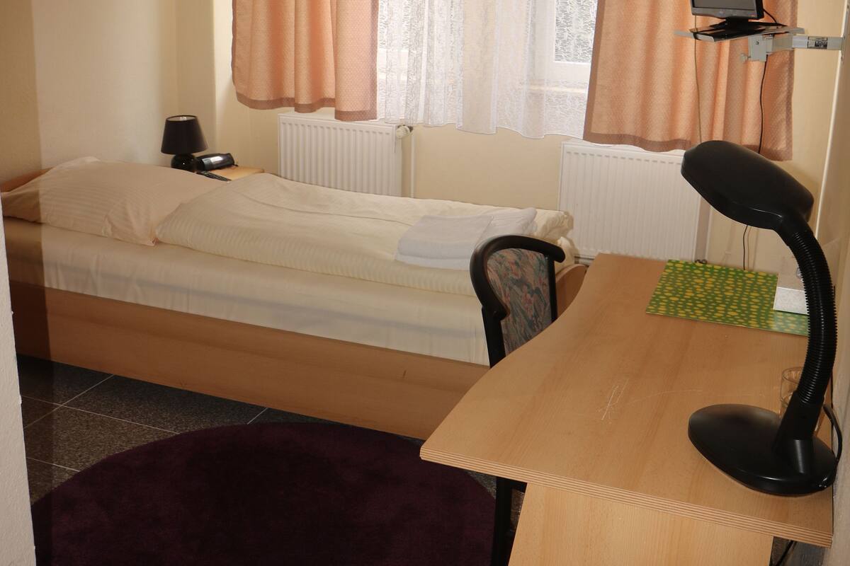 Ein kleines, ordentlich eingerichtetes Schlafzimmer mit einem Einzelbett, einem Schreibtisch mit Computer und einem Fenster mit Vorhängen.