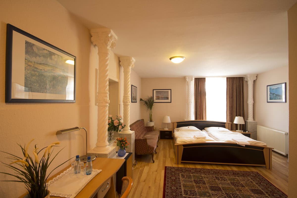 Ein Hotelzimmer mit Bett, Nachttischen, Schreibtisch, Stuhl, Schreibtischlampe, einem Blumentopf und einem Fenster mit Vorhängen.
