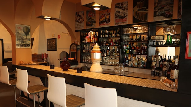 Zona de bar con taburetes blancos, una barra con diversas botellas y vasos, y un jarrón blanco decorativo.