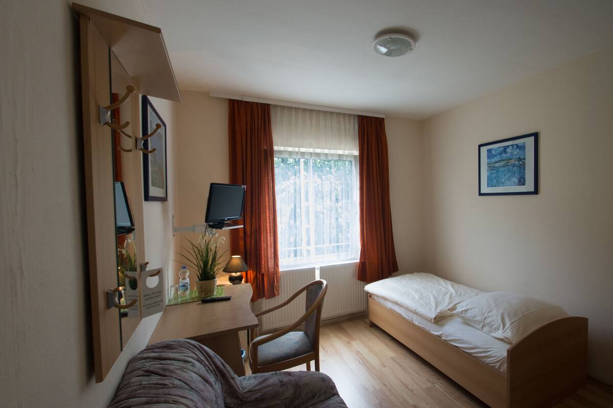 Ein gemütliches Zimmer mit Bett, Schreibtisch, Stuhl, Fernseher und einem fensterbelaubten Fenster.